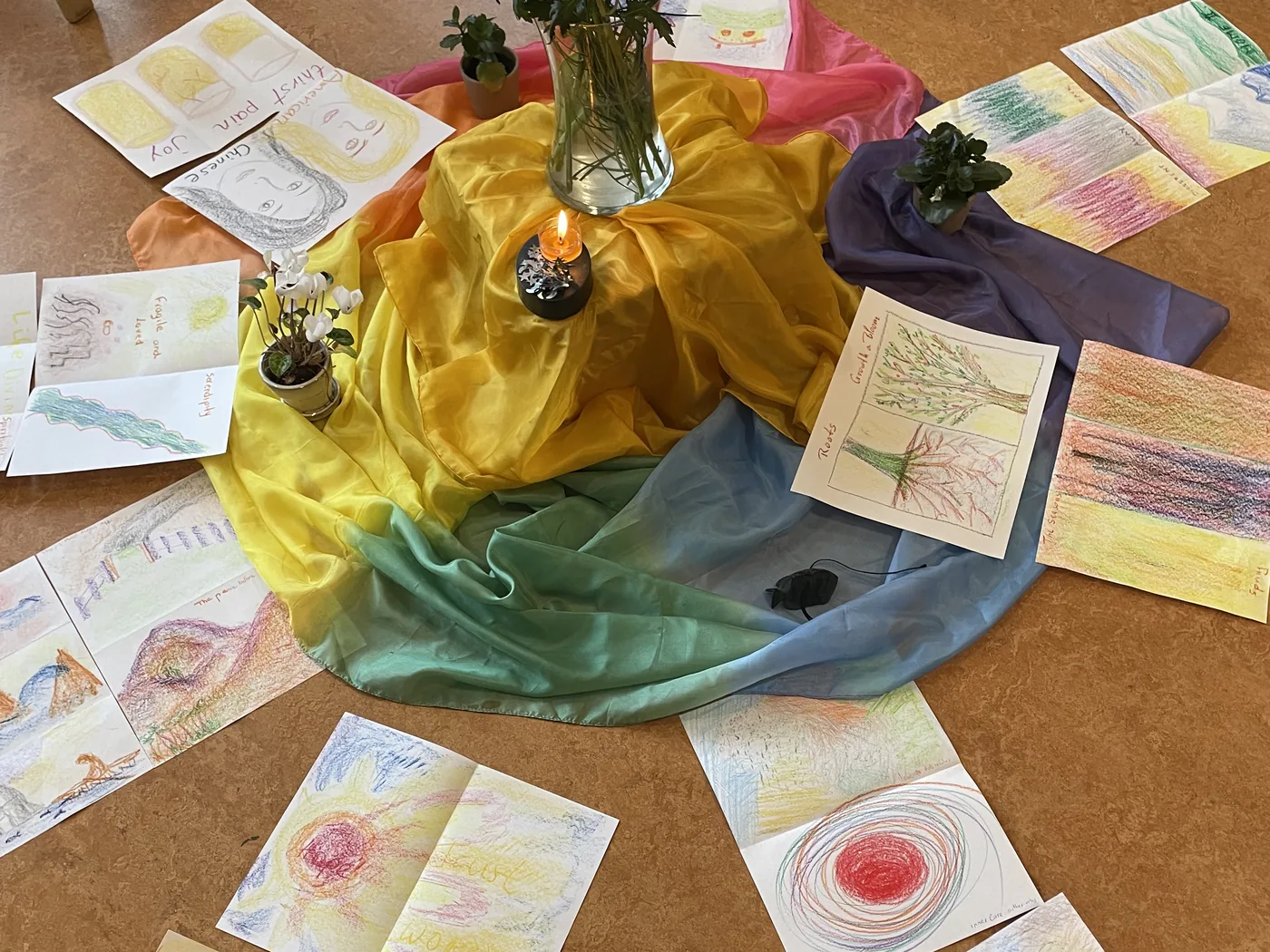 Trabajos en pastel y acuarela dispuestos en círculo alrededor de sedas de colores y flores